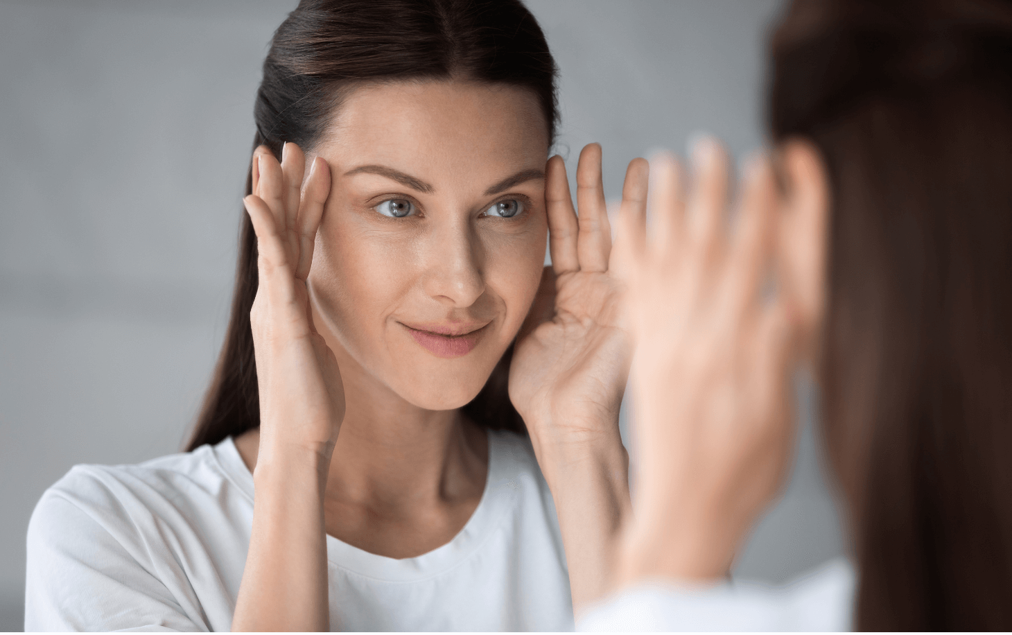 Woman with anti wrinkle treatment in Suffolk touching her cheekbones while looking in mirror smiling